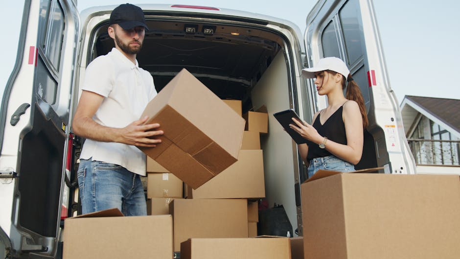 A man wearing a white polo shirt, blue jeans, and a black cap is inside the back of a white moving van, holding a large cardboard box as part of a home relocation process. The van is open, revealing several other cardboard boxes of various sizes stacked inside, some placed directly on the van floor and others on top of each other. Nearby, a woman dressed in a black tank top, denim shorts, and a white cap is standing outside the van, using a tablet device, likely coordinating the furniture transport or packing and moving tasks. The setting appears to be outdoors during daytime, with elements of a residential area visible in the background, including a partial view of a house. The scene captures a loading process that involves careful handling of packing materials, essential to efficient removals and reducing delays during house moves, as handled by companies like Ilford Removals.