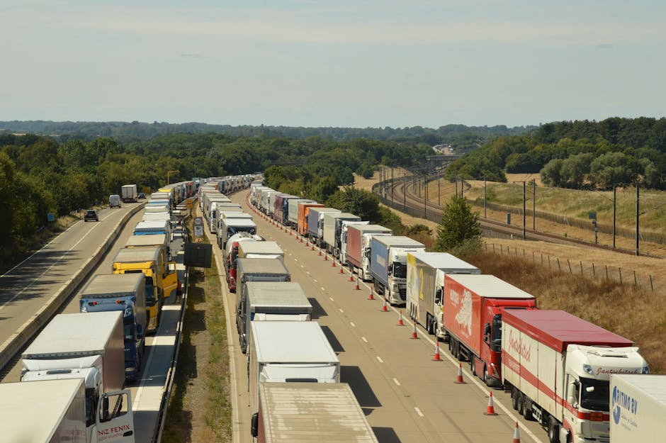 A long queue of trucks and lorries parked on the side of a two-lane road in a rural area, with some vehicles stretching into the distance. The trucks are arranged in a line, with some adjacent to the other lane, which appears to be partially blocked by additional vehicles and roadside cones. The trucks are used for furniture transport and house removals, with visible cargo areas covered by various coloured tarpaulins and wrapping materials. On the right side of the image, there is a grassy field with trees and a railway line running parallel to the road, while on the left side, a partly visible vehicle can be seen on the adjacent carriageway. The overall scene suggests a delay or congestion in the transportation of furniture and household items, which can occur during home relocations involving professional removals services, such as those offered by Ilford Removals. The background shows a distant horizon with low hills and a cloudy sky, indicating typical countryside surroundings for a moving operation.