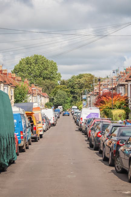 A residential street scene during daytime showing a row of parked cars on both sides of the road, including various models and colours of vehicles. In the background, there are terraced houses with brick façades, some with small front gardens and hedges, under a partly cloudy sky with overhead power lines visible. The street appears calm and typical of suburban urban areas, suggesting a suitable environment for house removals and home relocation services. Ilford Removals, a professional moving company, often operates in such neighbourhoods, providing furniture transport, packing, and loading services as part of comprehensive house removal and moving logistics. The scene captures the quiet, residential setting where furniture and boxes might be loaded onto a moving van as part of an organized home relocation process, with the focus on precision and professionalism in moving operations.