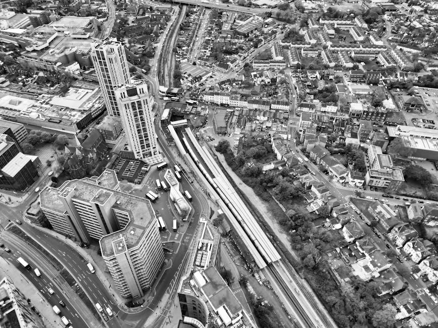 A black and white aerial photograph of an urban area showing tall office and residential buildings, with a railway station in the center featuring multiple platforms and trains. Surrounding the station are roads with various vehicles, including buses and cars, and a network of smaller streets lined with houses, shops, and greenery. The scene also includes a large railway bridge crossing over a wide road, with buses and cars visible in the traffic. The image captures a busy cityscape, likely representing part of Ilford, with dense development and transport infrastructure. The photograph illustrates an environment where house removals and furniture transport might take place, highlighting the complexity of urban logistics involved in relocation services provided by companies like Ilford Removals.