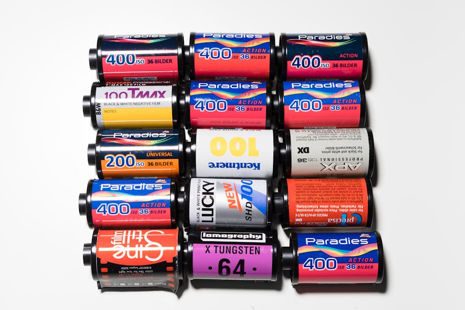 An overhead view of fourteen rolls of photographic film and photographic paper film rolls arranged in three rows on a white background. The top row contains three rolls of Paradies 400 ISO film, each with red and blue labels, positioned side by side. The middle row features five rolls, including a yellow-labeled roll of 100 Tmax black and white negative film, a white roll of Keneric 100, two more Paradies 400 ISO film rolls with red and blue labels, and a black and orange roll of 35mm film. The bottom row has six rolls, including an orange roll of 35mm film, a black roll of Tungsten X film, a purple roll of Fuji X Tungsten, and three more Paradies 400 ISO film rolls. The rolls are in close proximity, illustrating the packaging materials used for photographic and imaging supplies. This arrangement is related to moving and packing services by Ilford Removals, emphasizing careful handling of fragile and valuable items during home relocation or furniture transport processes.