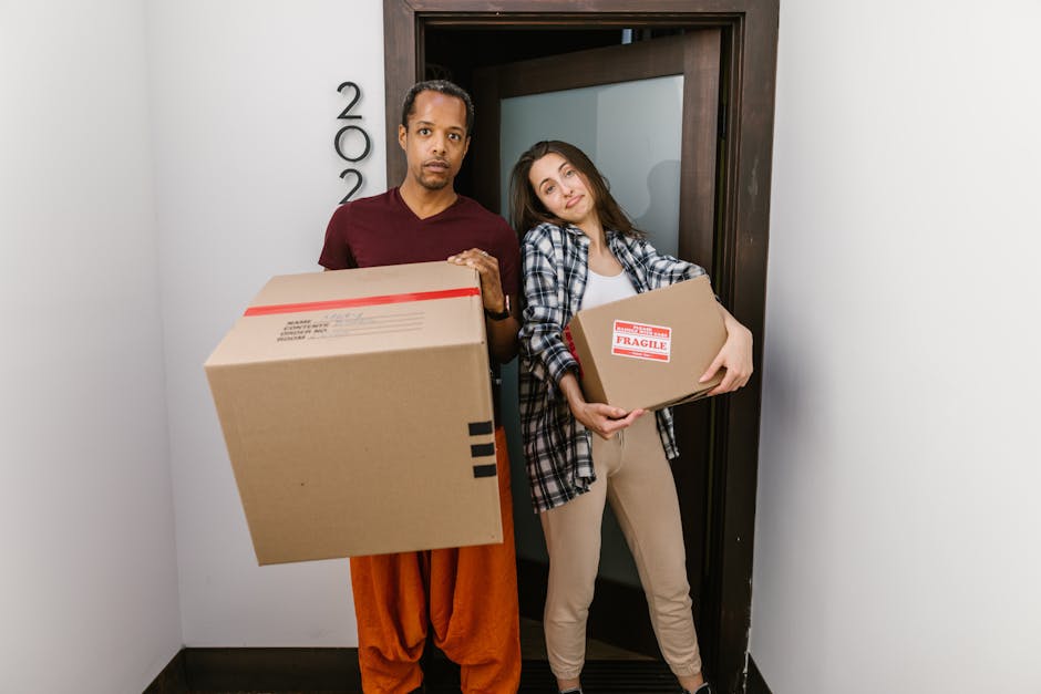 Two individuals, a man and a woman, are standing inside a building at an entrance door with the number '200' visible on the wall beside them. The man, dressed in a maroon shirt and orange trousers, is holding a large cardboard box with red and black labels, indicating it contains fragile items, and appears to be preparing for a home relocation. The woman, wearing a plaid shirt and beige trousers, is holding a smaller cardboard box with similar labels and is leaning her head slightly towards the man, suggesting a team effort in the furniture transport process. The entrance area has plain white walls and dark flooring, with natural lighting illuminating the scene. This image reflects the process of packing and loading boxes during a house move, with the involvement of professional removals, such as those offered by Ilford Removals, supporting logistics for tight access moves during a home relocation.