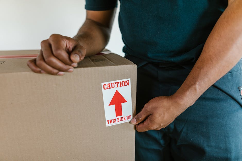 Close-up of a person wearing a dark teal shirt and dark trousers, holding a large cardboard moving box with both hands, one hand gripping the side and the other supporting the top, inside a residential property during a home relocation. The box has a prominent red and white caution label with an upward-pointing arrow and the words 'CAUTION THIS SIDE UP' to indicate proper orientation for safe handling. The background shows part of the interior doorway and floor, suggesting the loading process is occurring either inside a home or close to an entryway, with natural lighting illuminating the scene. This image illustrates the careful packing and handling stages involved in furniture transport and moving services as provided by Ilford Removals, highlighting attention to detail during packing and the importance of correct box orientation during the loading process for tight access moves near Ilford station.