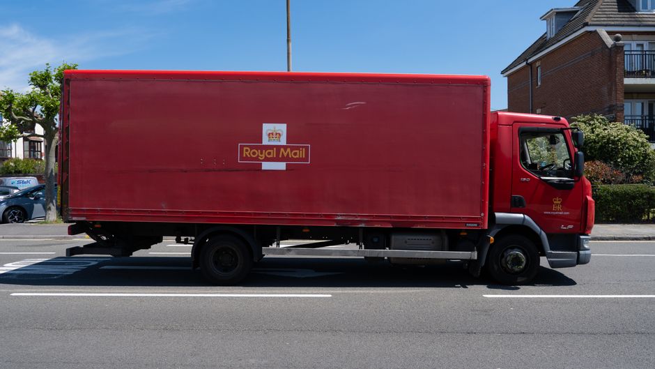 A red Royal Mail delivery truck parked in a residential area showing houses with pitched roofs and dormer windows, and green bushes along the pavement. The truck is positioned parallel to the curb and