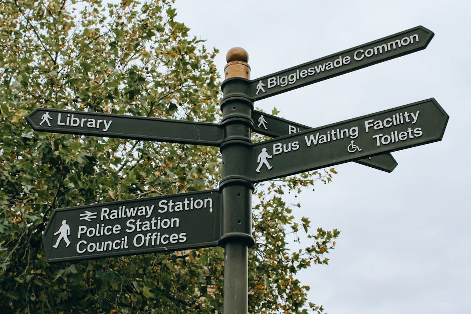 A black multi-directional street signpost situated outdoors against a background of leafy trees and a cloudy sky. The signpost features multiple rectangular signs with white text and symbols indicating directions to various locations including Biggleswade Common, Library, Railway Station, Police Station, Council Offices, Bus Waiting Facility, and Toilets. Some signs include pedestrian icons, and others display symbols such as a wheelchair for accessible facilities. The signpost is aligned on a central pole, with each sign extending outward in different directions, assisting residents and visitors in navigating the area. The scene appears to be in a town or urban setting, relevant to local house removals, relocation planning, and moving logistics, as facilitated by companies like Ilford Removals, supporting efficient furniture transport and packing during home relocation.