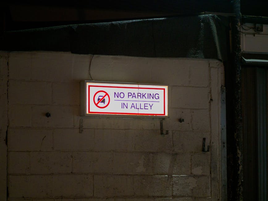 Illuminated rectangular sign mounted on a beige brick wall at night, displaying the message 'NO PARKING IN ALLEY' in blue uppercase letters. The sign features a red circle with a diagonal line crossing out a black arrow pointing downward, indicating parking restrictions. It is positioned above a doorway or garage entrance, with dark shadowed areas surrounding it. This signage is relevant for house removals and furniture transport, as it indicates a parking restriction that may affect loading and unloading processes during home relocation activities, with the sign being part of the urban environment managed by Redbridge Council, in Ilford. Ilford Removals sometimes operates within such regulated areas to coordinate efficient removal services and comply with local parking rules for removals.