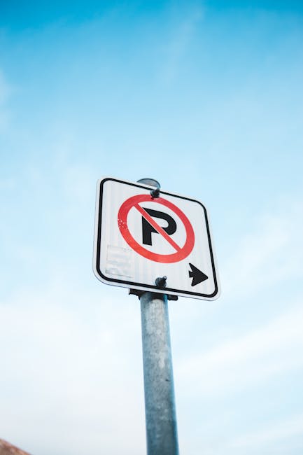 A black multi-directional street signpost situated outdoors against a background of leafy trees and a cloudy sky. The signpost features multiple rectangular signs with white text and symbols indicating directions to various locations including Biggleswade Common, Library, Railway Station, Police Station, Council Offices, Bus Waiting Facility, and Toilets. Some signs include pedestrian icons, and others display symbols such as a wheelchair for accessible facilities. The signpost is aligned on a central pole, with each sign extending outward in different directions, assisting residents and visitors in navigating the area. The scene appears to be in a town or urban setting, relevant to local house removals, relocation planning, and moving logistics, as facilitated by companies like Ilford Removals, supporting efficient furniture transport and packing during home relocation.