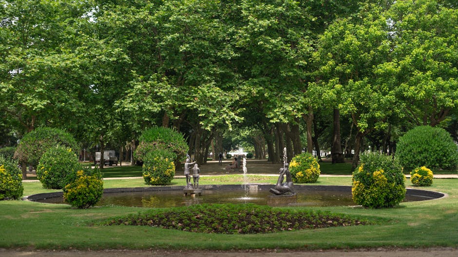 A scenic park setting featuring a circular fountain with multiple small sculptures of children and animals, surrounded by neatly trimmed bushes and vibrant green grass, with a central pond area that includes flowing water and decorative statues. Large trees with lush, dense foliage provide shade and create a canopy over the park, extending into the background where more greenery and a pathway are visible. The scene depicts a peaceful environment ideal for home relocation or outdoor activities, with the park's landscaping emphasizing organized planting and aesthetic arrangement suitable for house removals and transport logistics, as seen through the visual cues of carefully maintained vegetation and open space.