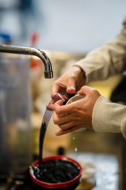 A person wearing a beige long-sleeved shirt is holding a metal measuring tape with both hands, examining it closely in a workspace. In the background, there are blurred objects including a large stainless-steel sink or container with a faucet, and various tools and materials suggesting a preparation or packing area for house removals. The setting appears to be indoors with sufficient lighting, possibly within a storage or packing facility used by Ilford Removals for home relocation and furniture transport tasks. The individual’s focus on the measuring tape indicates measurement or packing activities related to the logistics of loading and unloading furniture and boxes during a move.