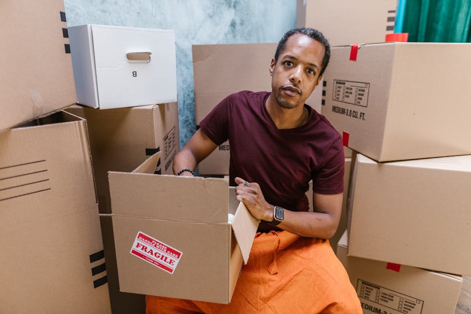 A man seated amid numerous cardboard boxes, some marked with a red 'Fragile' label, inside a room with a blue and orange wall in the background. He is wearing a maroon t-shirt, orange pants, and a watch, and he appears to be in the process of packing or unpacking. The boxes are of various sizes, stacked around him, with some open revealing packing materials such as paper and bubble wrap. The scene suggests a home relocation or furniture transport scenario, as part of a professional removals service, possibly provided by Ilford Removals, involving the careful handling and organization of household items during the loading process. The ambient lighting is neutral, highlighting the details of the packing activity and environment, which aligns with the logistics of house removals and moving services, focusing on safe packing and transportation of possessions during a move.